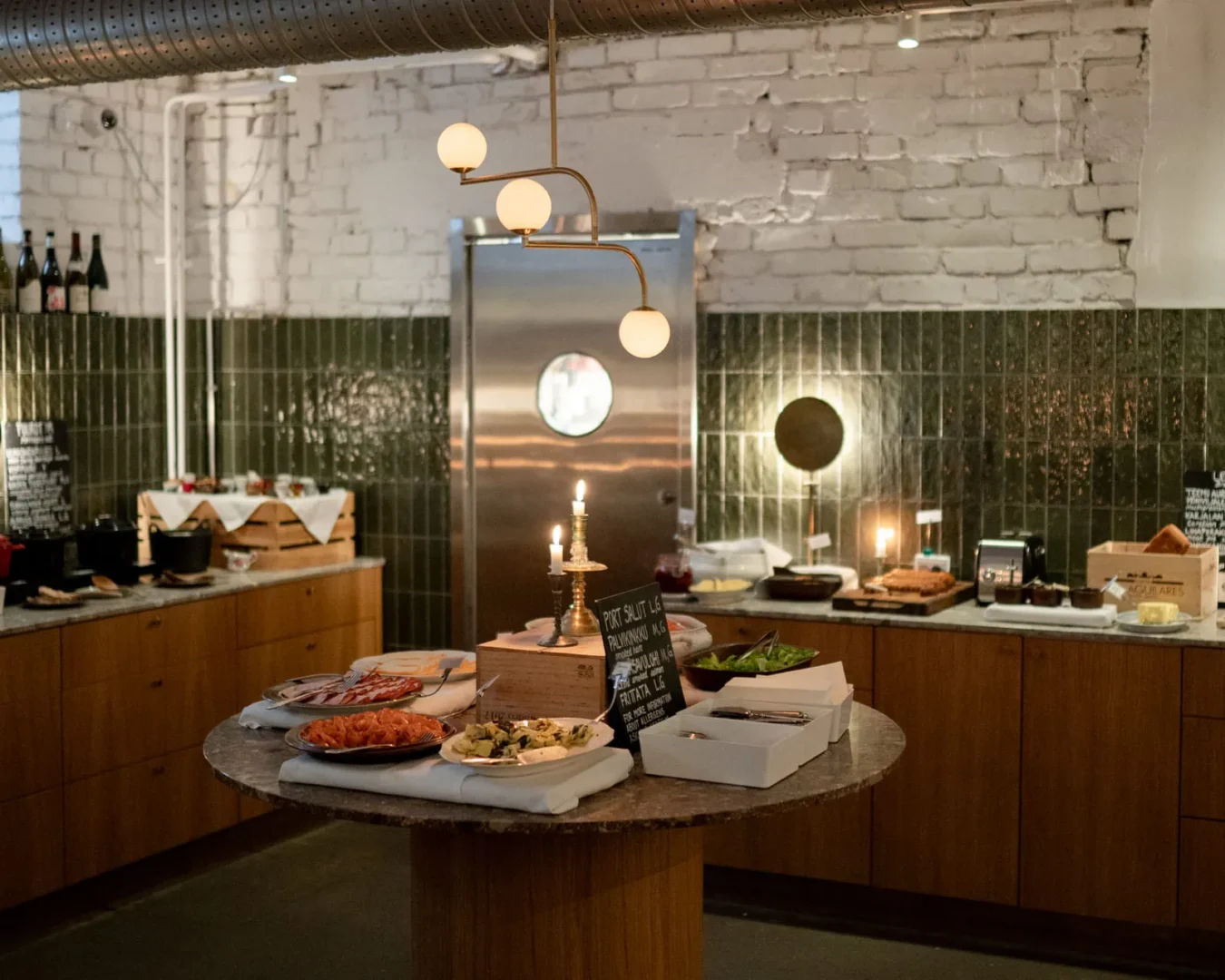 Round buffet table in a warm, industrial kitchen, with candles, plates of food, and a chalkboard menu centerpiece