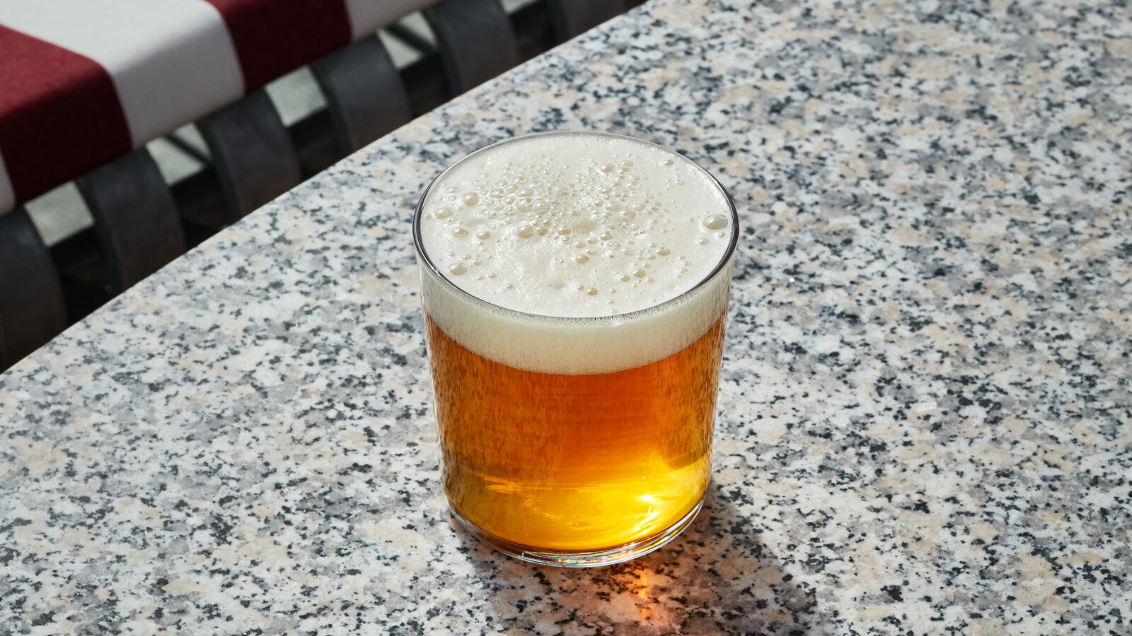 Pint of amber beer with frothy head on a speckled granite table outdoors, with a red-and-white striped umbrella in the background.