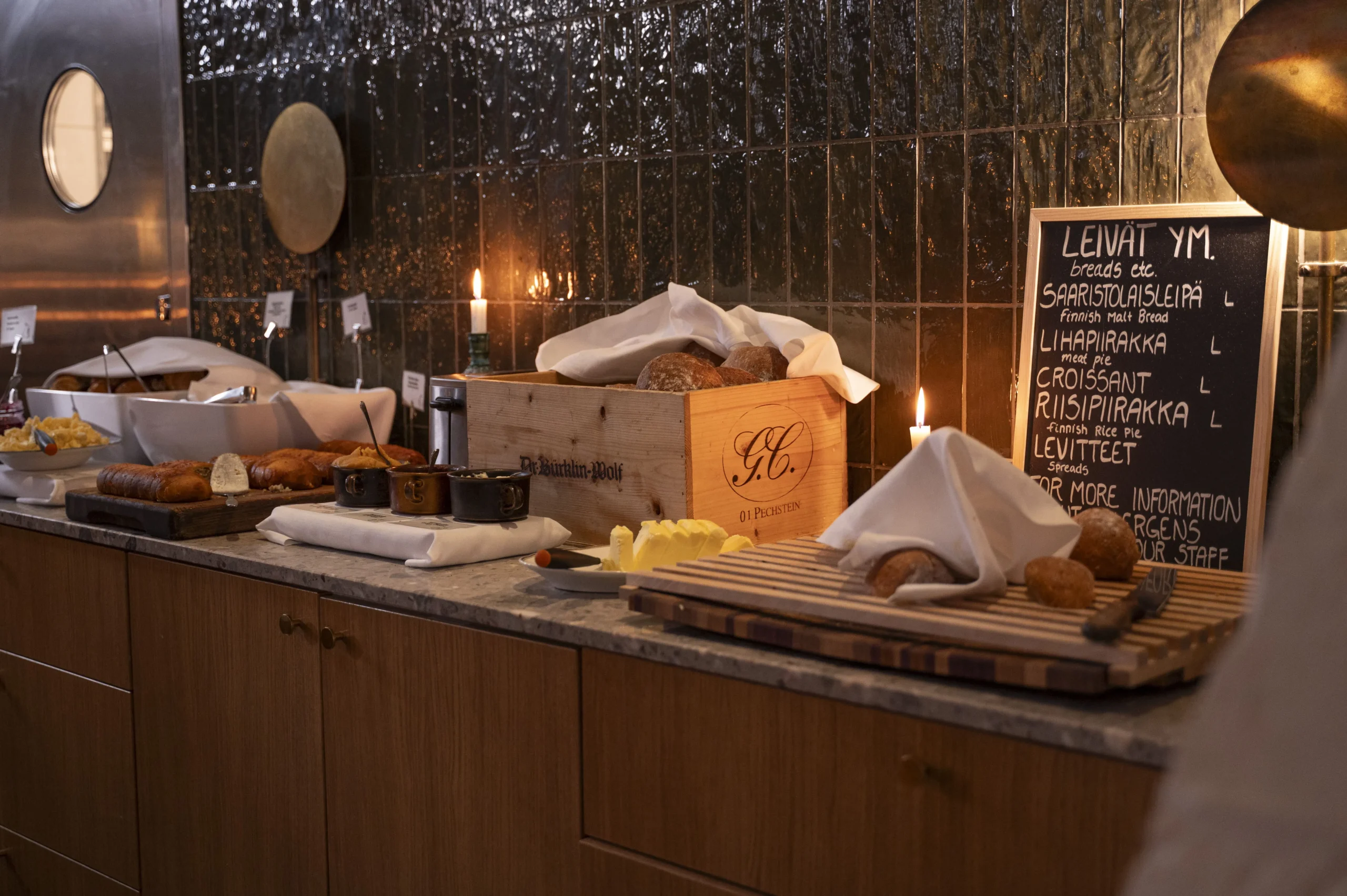 Bakery counter with assorted bread loaves in a wooden crate, butter, and a chalkboard menu beside warm candlelight.
