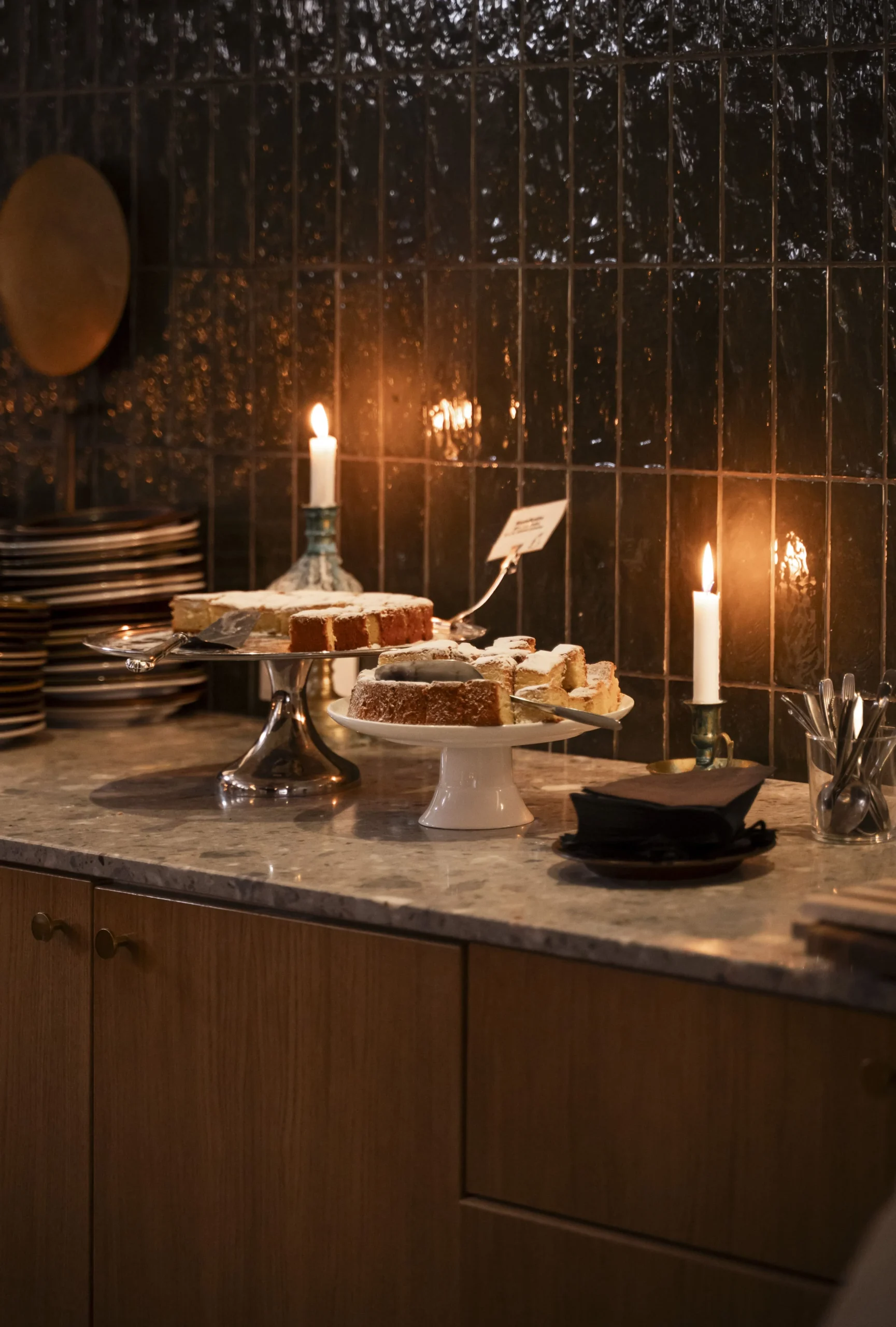 A marble kitchen counter with plates of cake on a white stand and lit candles in a dark tiled backsplash setting behind them.