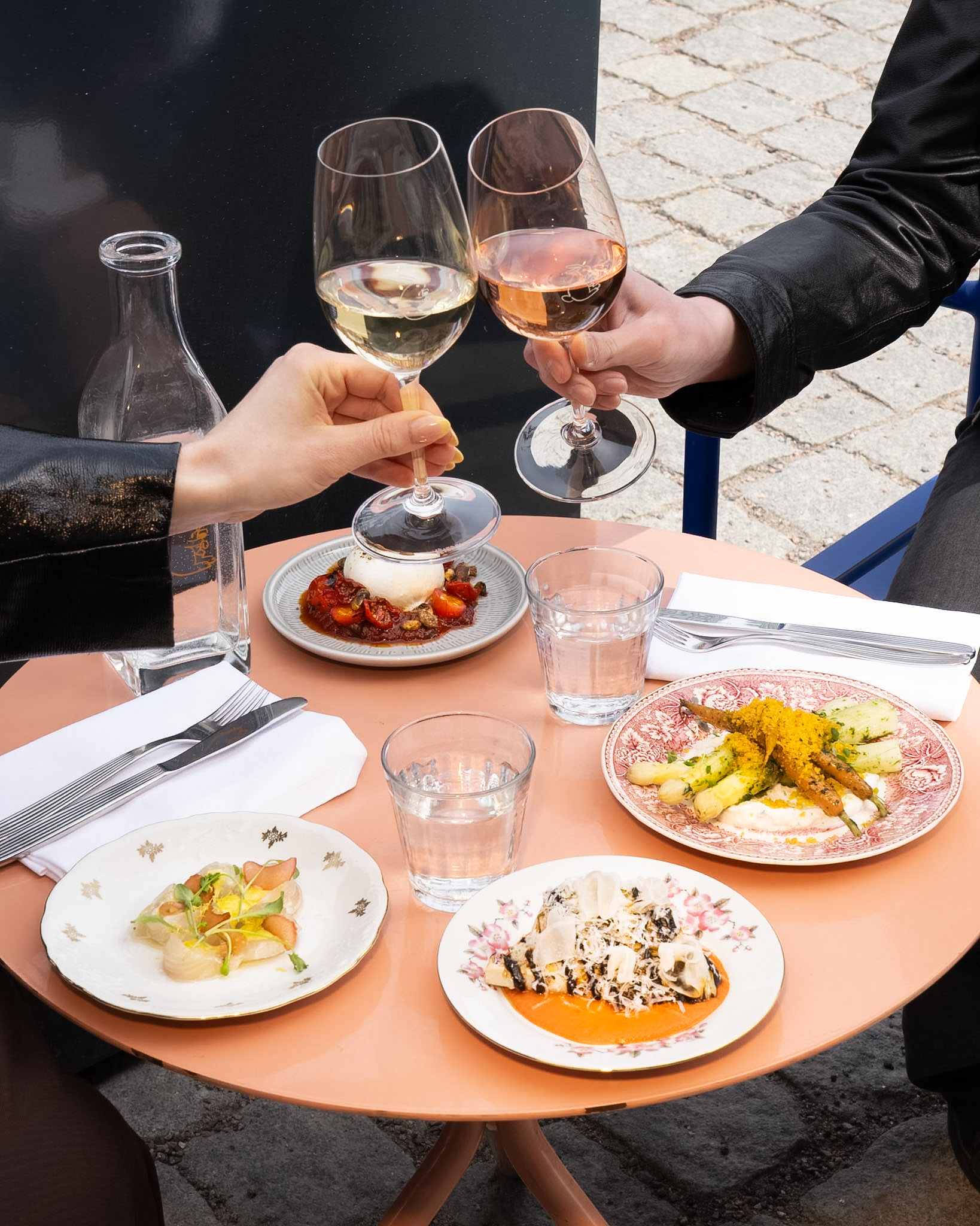People clinking wine glasses over a table with dishes and a pink table in an outdoor setting