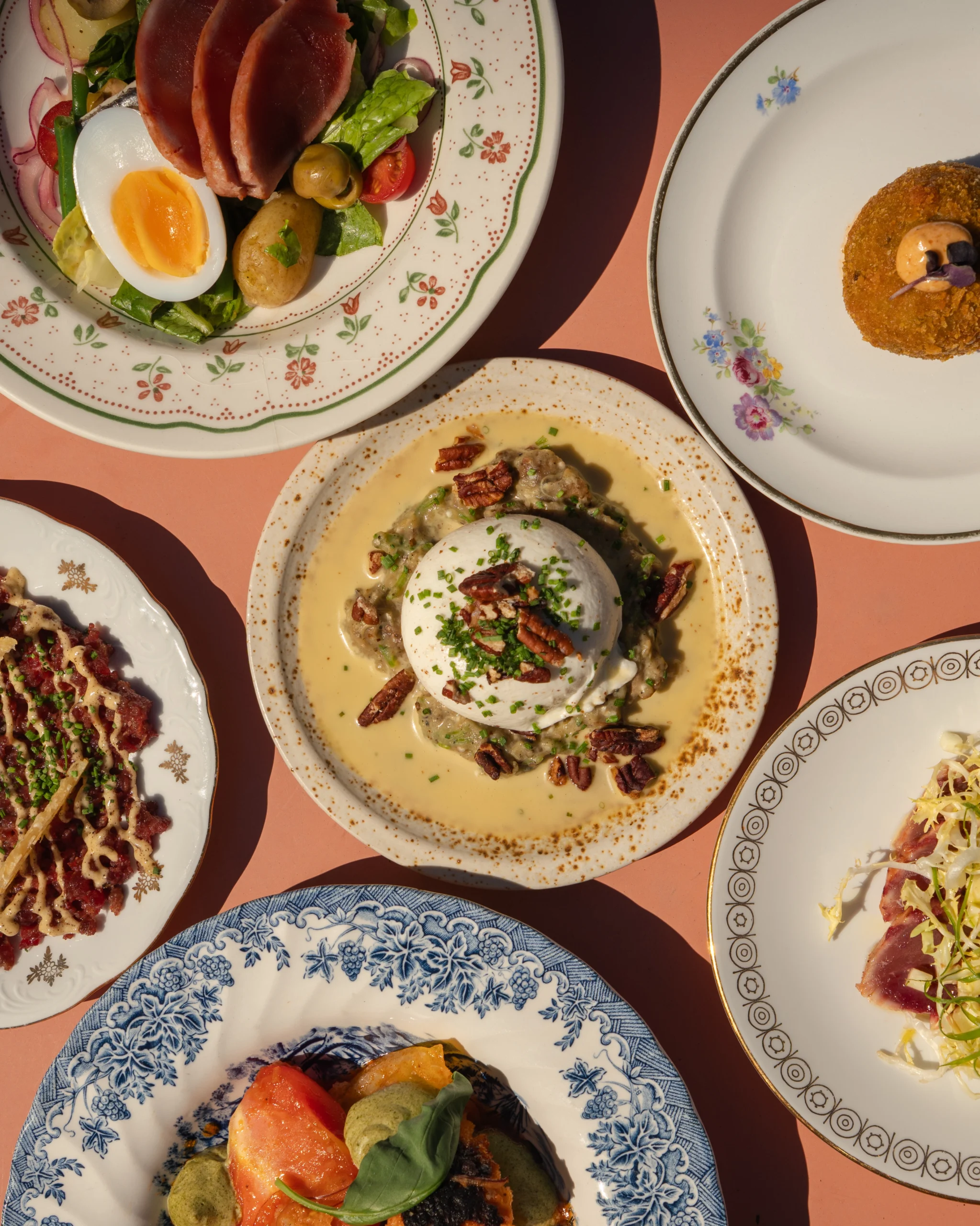 Assorted plates of food on a peach table, including a salad with boiled egg, olives, and slices of cured meat.