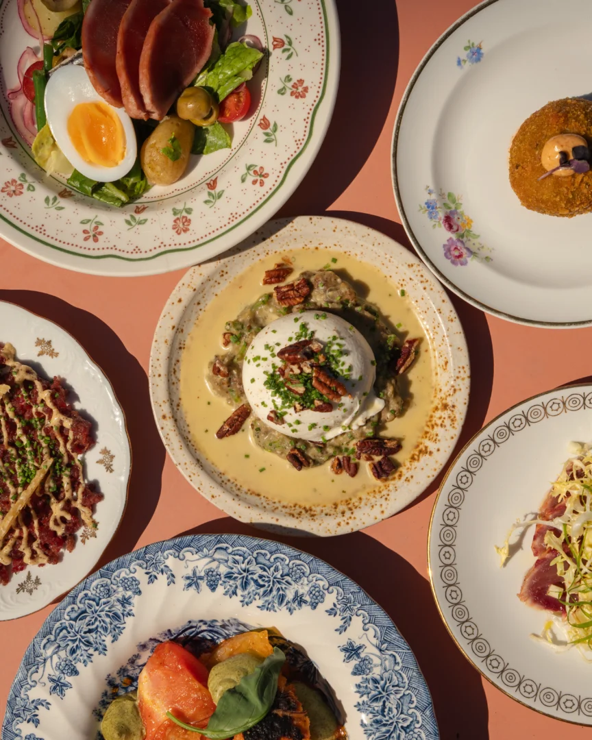Assorted plated dishes on a pink table, featuring a salad with a soft-boiled egg, greens, tomatoes, and sliced meat.