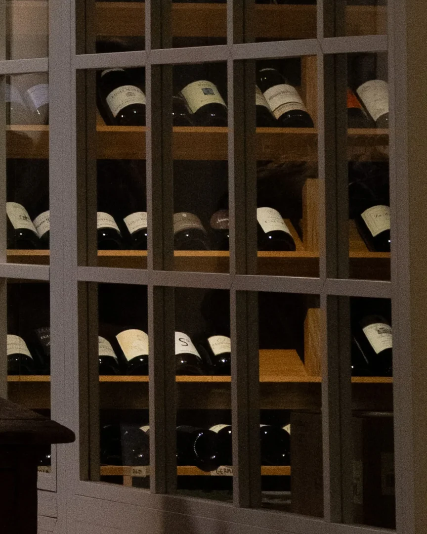 Rows of wine bottles stored on wooden shelves behind a glass-front cabinet in a dimly lit wine rack area