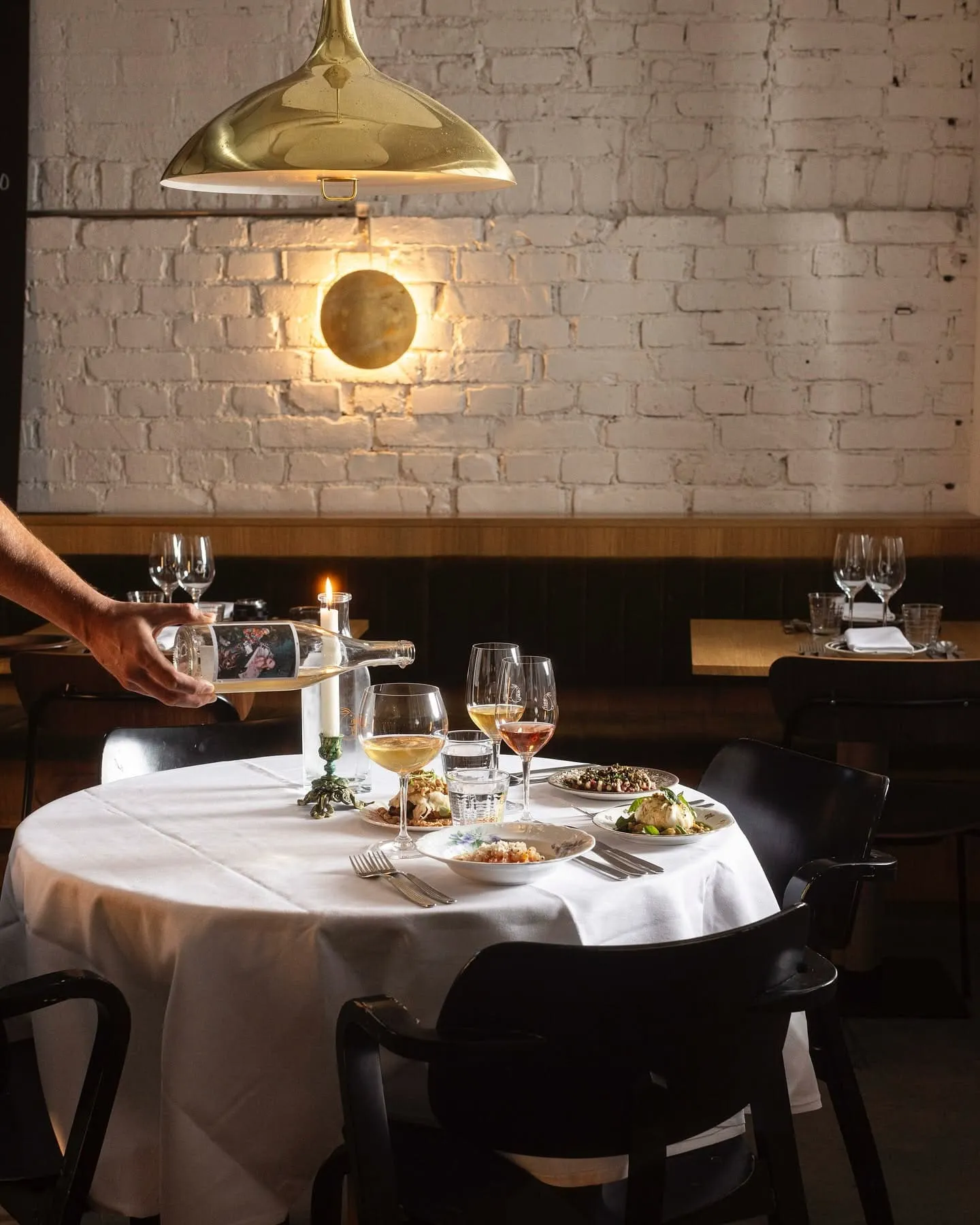 Waiter pours wine at a candlelit table set for two in a restaurant with a white brick wall backdrop.