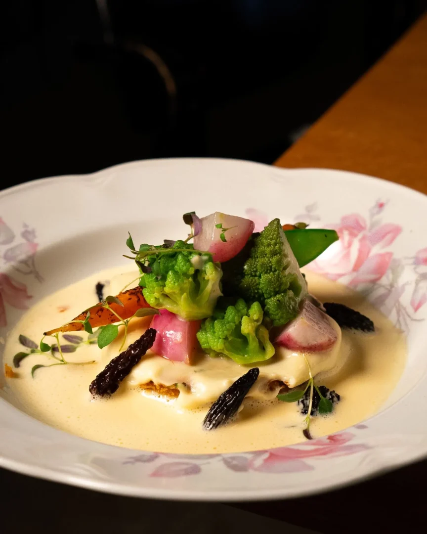 Close-up of a plated gourmet dish with pink radish, green broccoli, herbs, and a pale creamy sauce with dark crisp garnish on a floral plate.