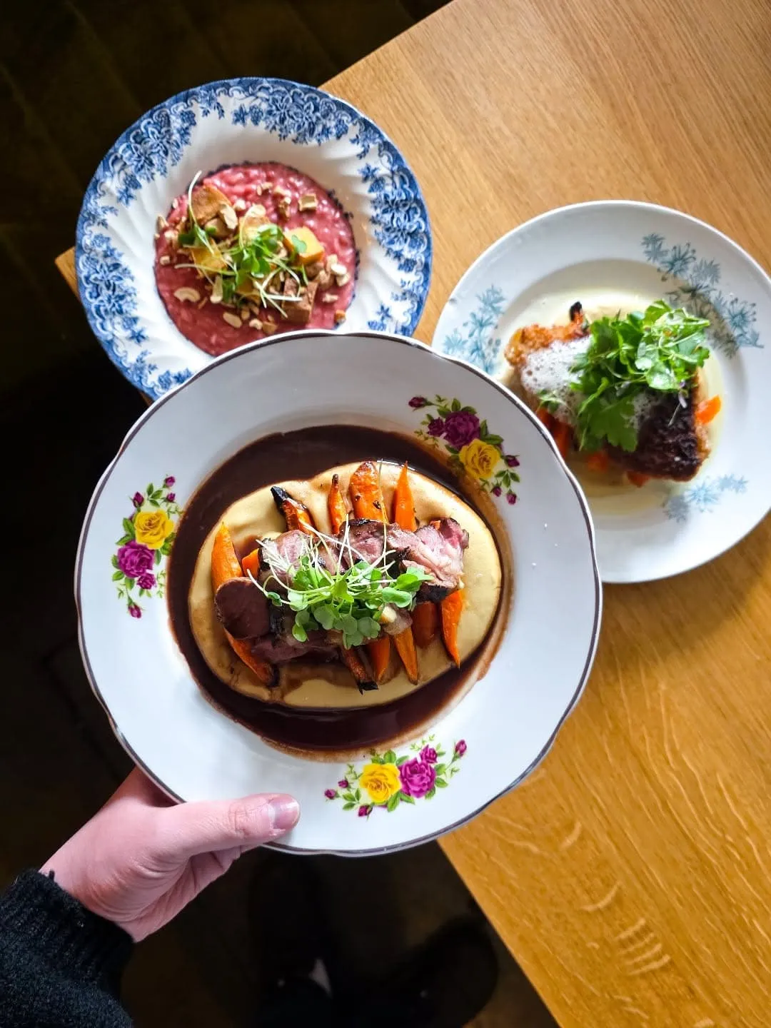 Overhead view of three plated dishes on a wooden table: center plate with beef, carrots, and creamy purée, topped with microgreens.