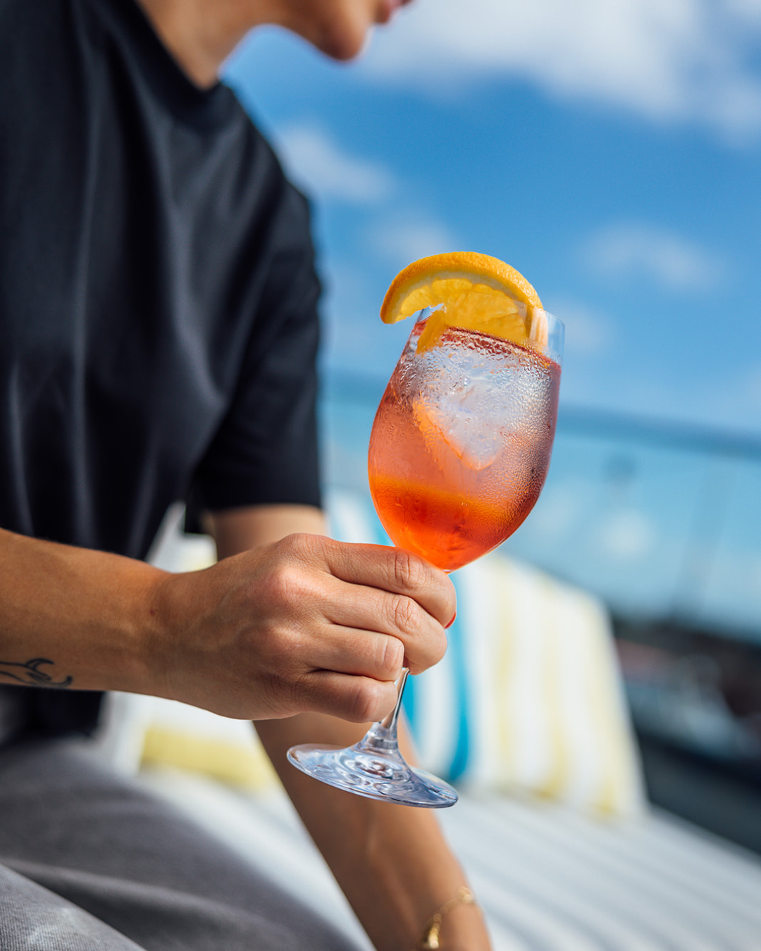 A person in a black shirt holds a stemmed cocktail glass with an orange slice garnish on a sunny deck by the water.