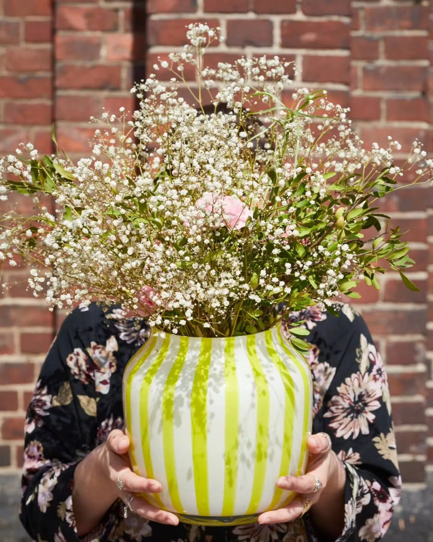 Person in a floral outfit holds a lime-green striped vase overflowing with white baby's breath flowers against a red brick wall.