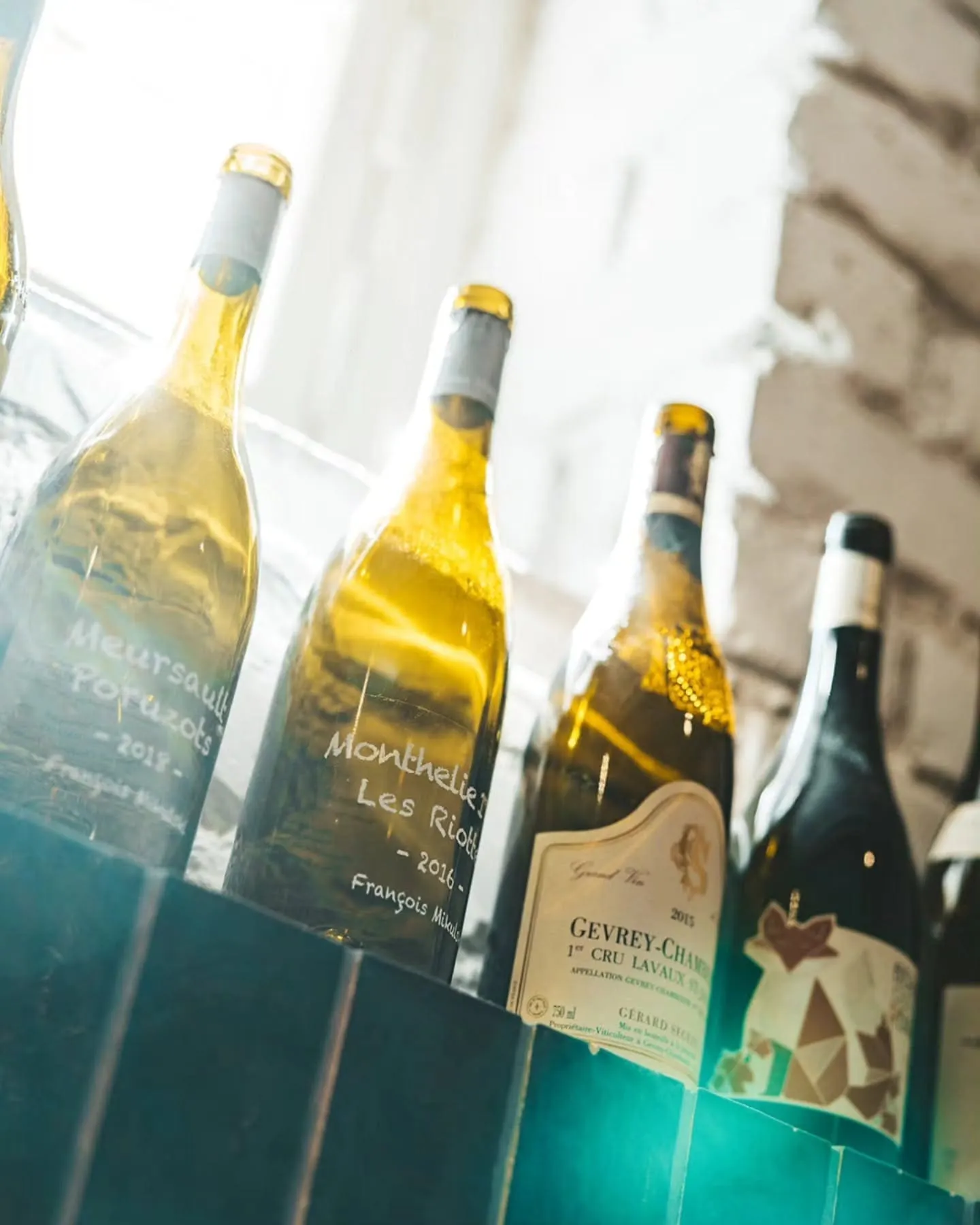 Several wine bottles tilted in a glass refrigerated display, labels partly legible (e.g., Gevrey-Chambertin) with gold foil tops in the foreground.